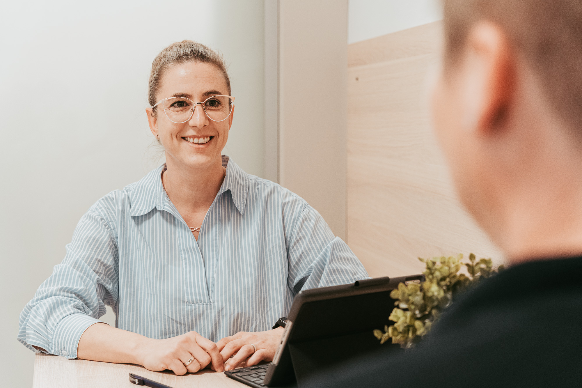 Eine lächelnde Frau mit Brille sitzt an einem Tisch, vor ihr ein Tablet.